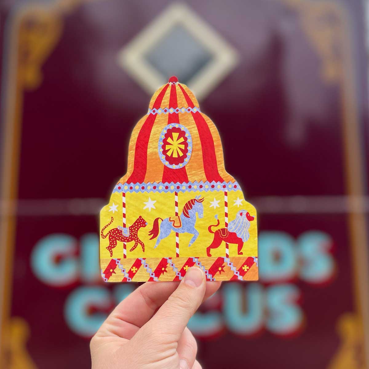 Carousel Shaped Greeting Card5 Kartka w kształcie karuzeli, ozdobiona kolorowymi końmi i lwem na tle ciepłych barw. Ilustracja ma piękny retro klimat. Wydrukowana w Wielkiej Brytanii na papierze z recyklingu FSC. W zestawie znajduje się koperta.
